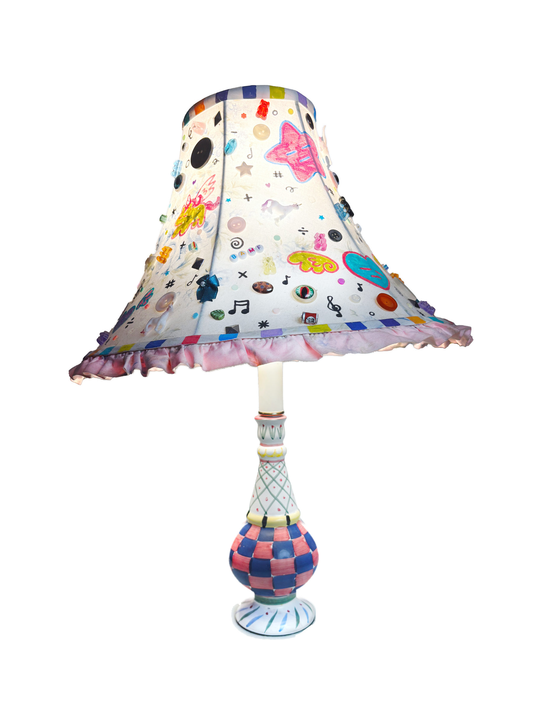 Bell-shaped lamp with a ruffled pink hem. The white shade is covered in drawn and attached motifs: buttons, stars, music notes and butterflies. The base is a slim vase with a pink and blue checked bulb.