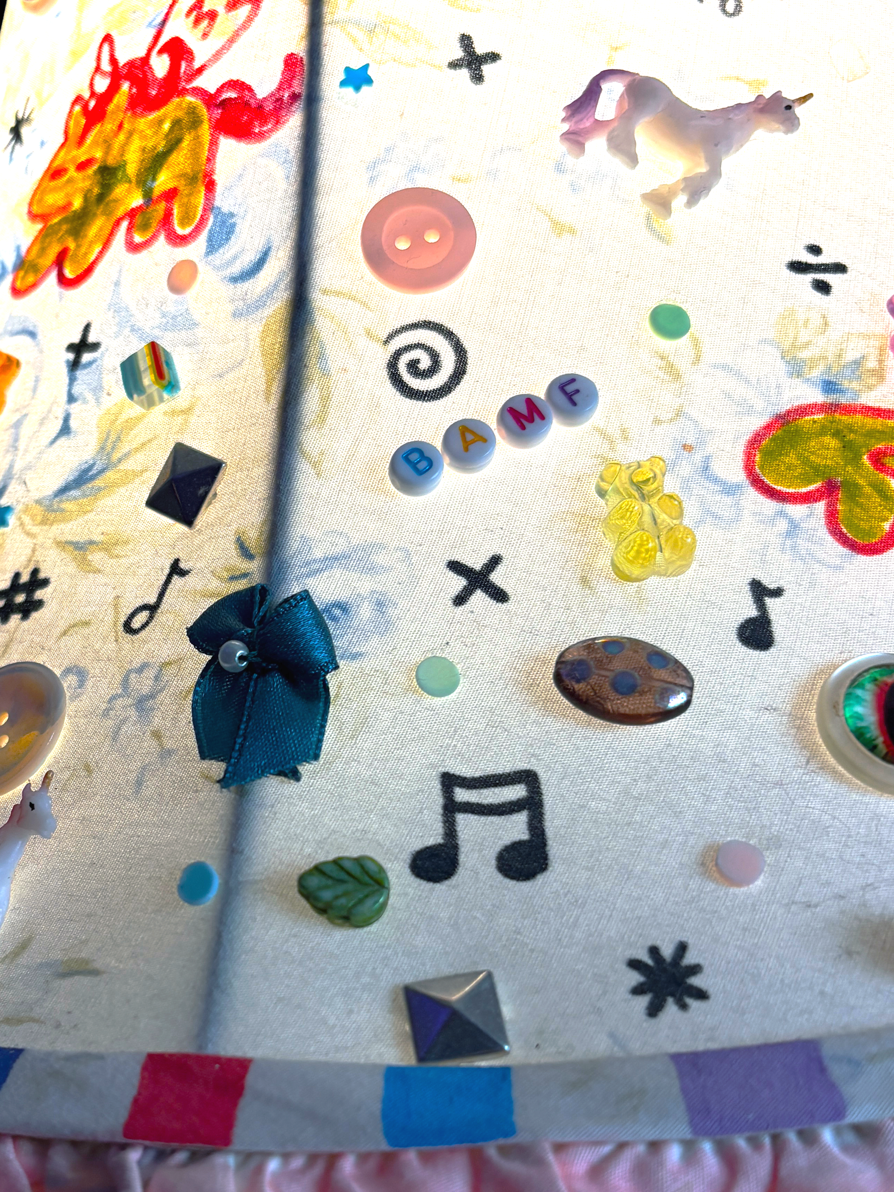 Close-up of the bell lamp's shade: buttons, a teal bow, a gummy bear and a unicorn charm among drawn stars and music notes.