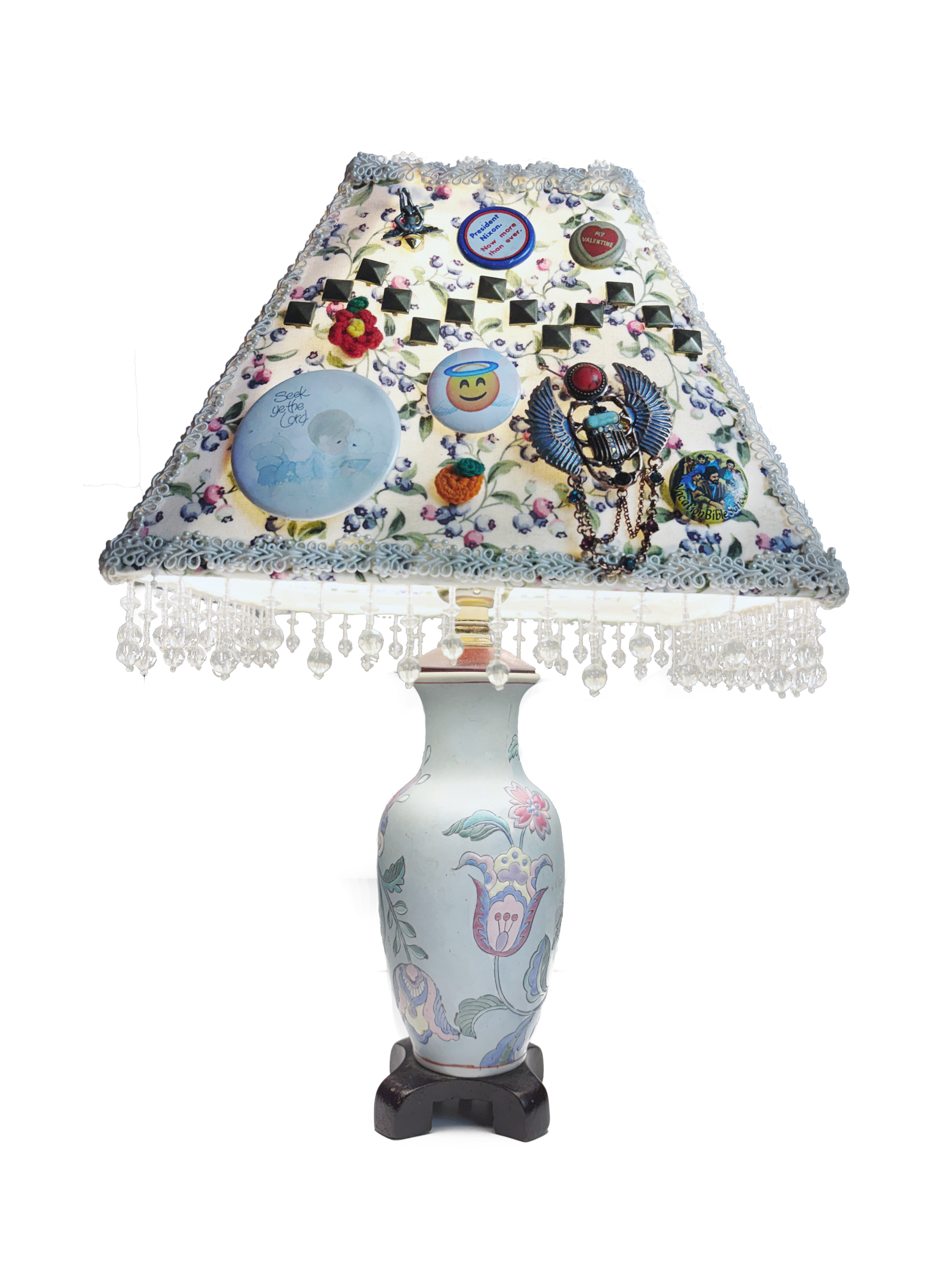 The same lamp from the fourth side, its shade covered in pinback buttons, a smiley face, a winged scarab charm and beaded chains.